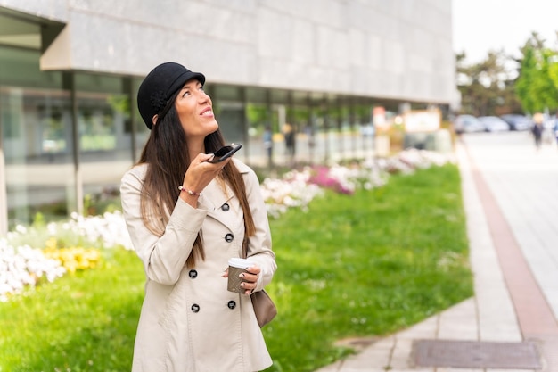 Imprenditrice fuori dall'ufficio con un caffè in mano e l'invio di una nota vocale con il telefono