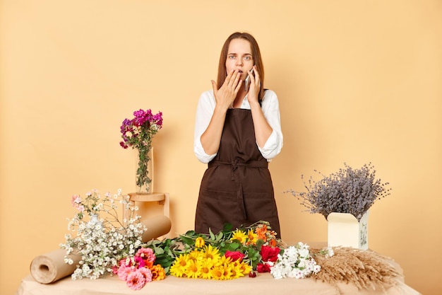 Imprenditrice scioccata e sbalordita che indossa un grembiule marrone in piedi nel proprio negozio di fiori e parla sullo smartphone con un cliente che posa isolato su uno sfondo beige