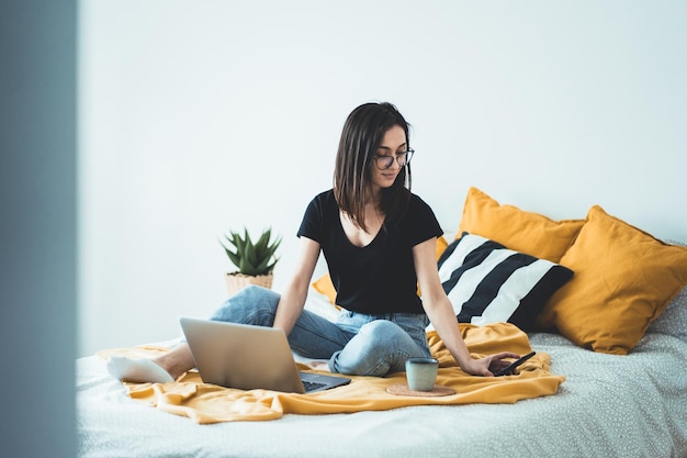 Imprenditrice seduta a letto con un portatile e che controlla lo smartphone mentre lavora a casa