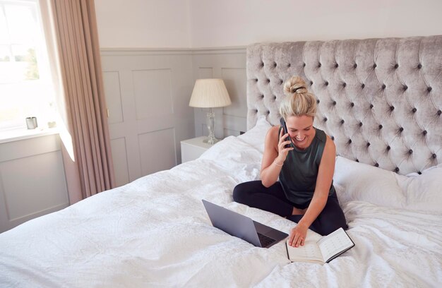 Imprenditrice seduta sul letto con laptop e telefono cellulare che lavora da casa durante il blocco pandemico