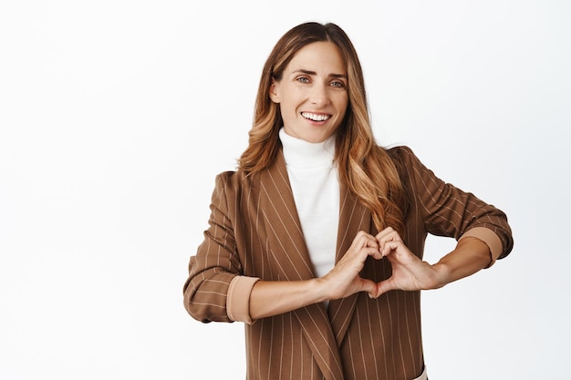 Imprenditrice sorridente che mostra il segno del cuore della mano in piedi in abito marrone su sfondo bianco