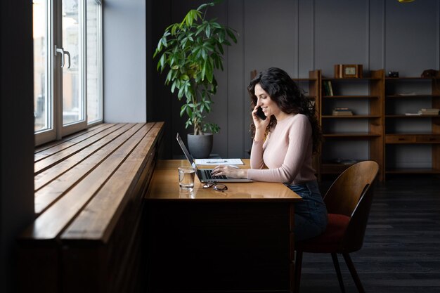 Imprenditrice sorridente che parla con il cliente tramite dispositivo mobile che lavora con l'ordine online sul laptop