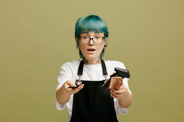 Impressionato giovane barbiere femminile che indossa occhiali uniforme guardando la fotocamera che allunga pettini e spray per capelli verso la fotocamera isolata su sfondo verde oliva