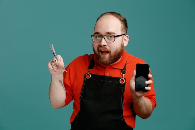 Impressionato giovane barbiere maschio con occhiali camicia rossa e grembiule da barbiere che mostra le forbici e il telefono cellulare guardando la fotocamera isolata su sfondo blu
