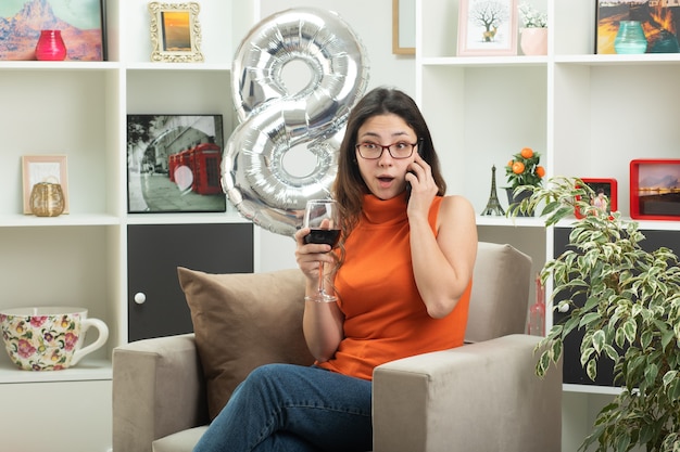 Impressionato giovane bella donna con gli occhiali parlando al telefono e tenendo un bicchiere di vino seduto sulla poltrona in soggiorno a marzo giornata internazionale della donna