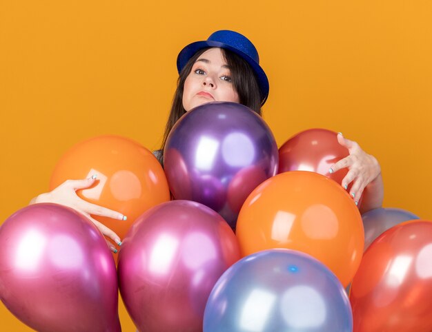 Impressionato giovane bella ragazza che indossa un cappello da festa in piedi dietro i palloncini