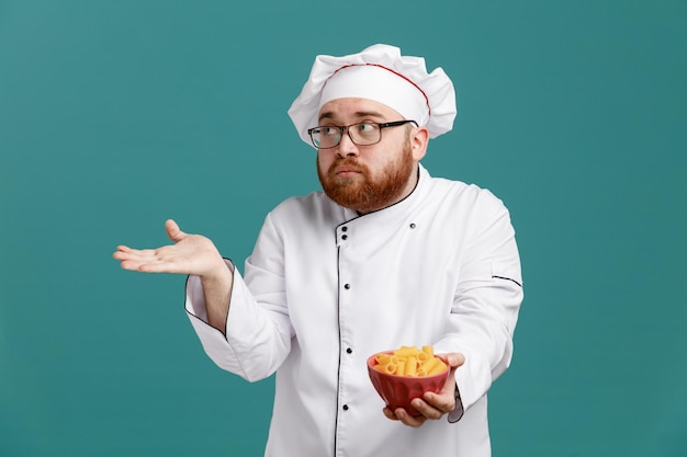 Impressionato giovane chef maschio che indossa occhiali uniformi e cappuccio che tiene una ciotola di maccheroni guardando il lato che mostra la mano vuota isolata su sfondo blu