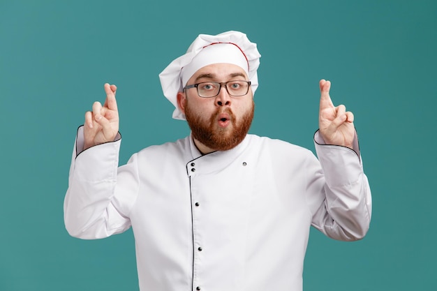 Impressionato giovane chef maschio che indossa occhiali uniformi e cappuccio facendo un gesto con le dita incrociate guardando la fotocamera isolata su sfondo blu