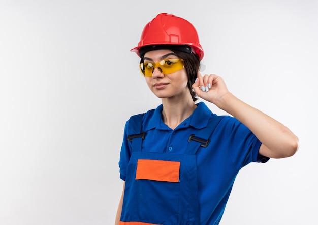 Impressionato giovane donna costruttore in uniforme con gli occhiali mettendo il dito sull'orecchio isolato sul muro bianco