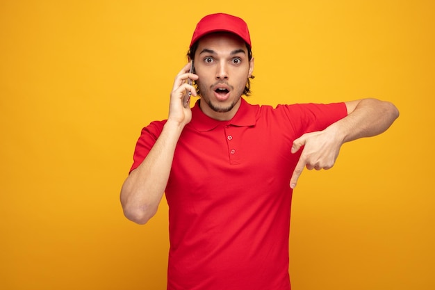 impressionato giovane fattorino che indossa uniforme e cappuccio guardando la fotocamera rivolta verso il basso mentre parla al telefono isolato su sfondo giallo