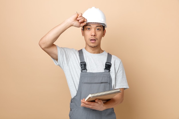 Impressionato giovane operaio edile che indossa il casco di sicurezza e uniforme che tiene la matita e il blocco note che afferra il casco