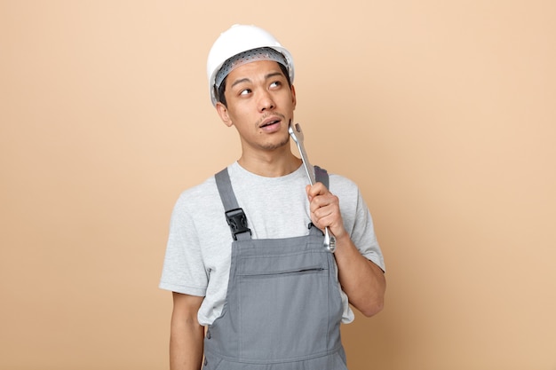 Impressionato giovane operaio edile che indossa il casco di sicurezza e uniforme che tocca la faccia con la chiave che osserva in su