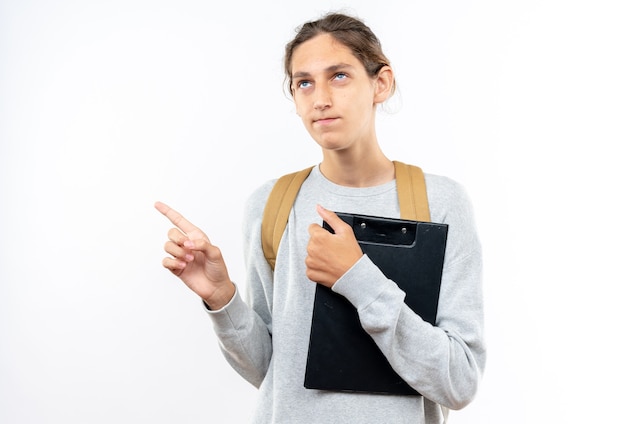 Impressionato giovane studente che indossa uno zaino che tiene i punti degli appunti sul lato isolato sul muro bianco con spazio di copia