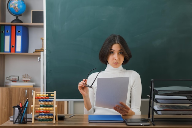 impressionato guardando la fotocamera giovane insegnante femminile che indossa occhiali tenendo la carta seduto alla scrivania con gli strumenti della scuola in classe