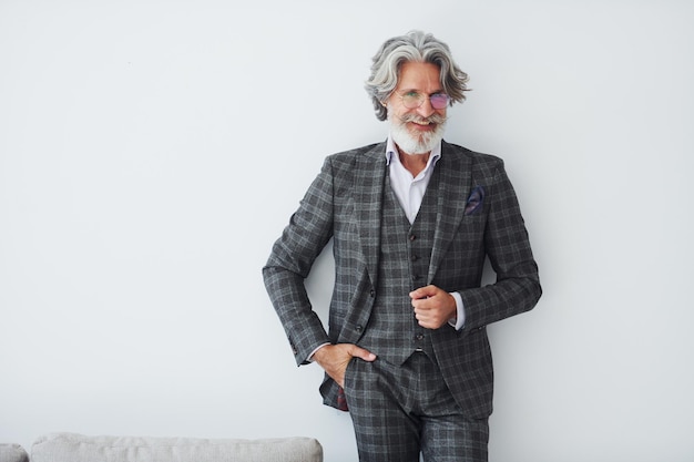 In abiti eleganti in piedi contro un muro bianco anziano elegante uomo moderno con capelli grigi e barba all'interno