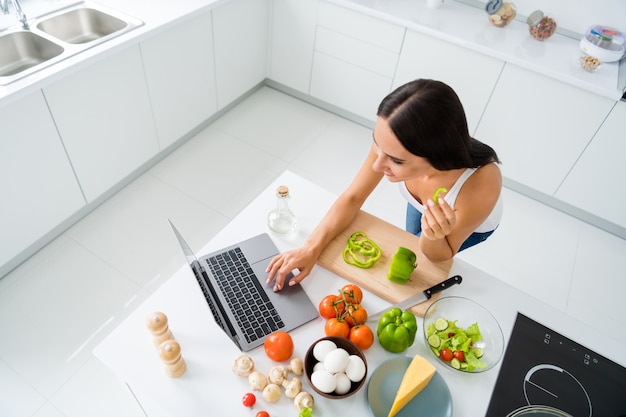 In alto sopra la foto ad alto angolo della casalinga positiva allegra vuole preparare la cena della festa usa il suo laptop cerca ricetta biologica vegetariana guarda film sulla cucina in cucina bianca casa al chiuso