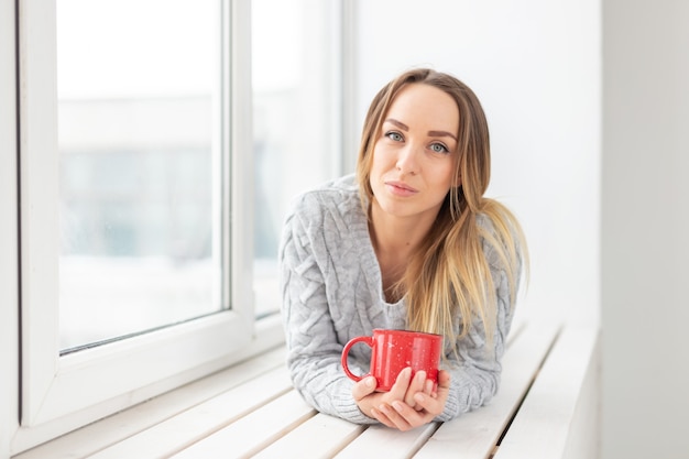 In attesa, solitudine e concetto di bellezza - giovane donna con una tazza di tè caldo seduto sul davanzale della finestra