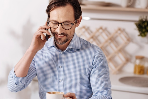 In contatto con tutti. Allegro felice barbuto lavoratore autonomo libero professionista seduto a casa e fare colazione mentre si conversa al telefono