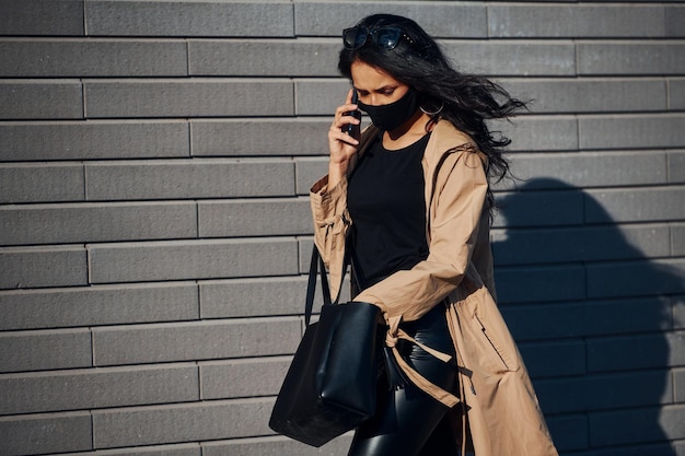 In maschera protettiva Bella bruna con capelli ricci e in abiti neri che parlano al telefono all'aperto vicino al muro