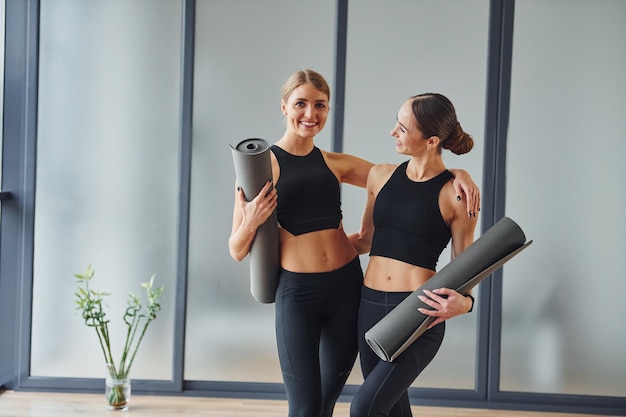 In piedi con le stuoie in mano Due donne in abbigliamento sportivo e con corpi snelli hanno una giornata di yoga fitness insieme al chiuso