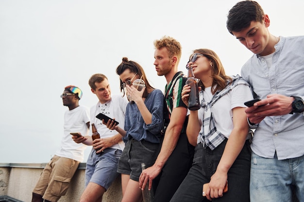 In piedi con telefoni e alcol in mano Gruppo di giovani in abiti casual organizzano una festa sul tetto insieme durante il giorno