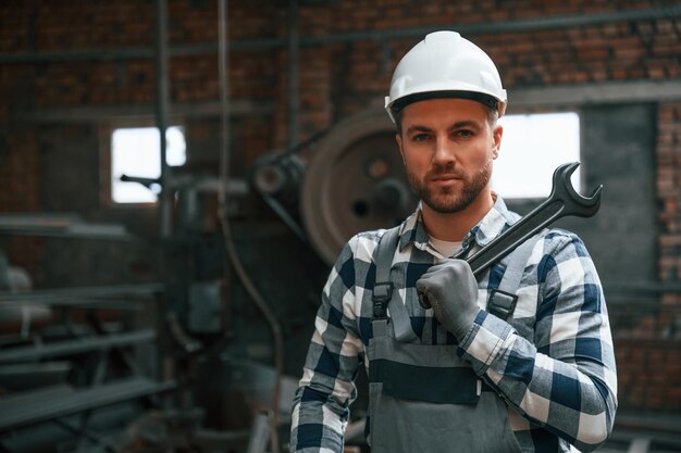 In piedi in uniforme grigia e con elmetto bianco Il lavoratore di fabbrica è al chiuso