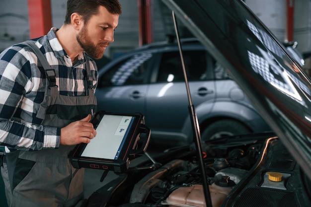 In piedi vicino all'auto con il cofano aperto Holding tablet L'uomo in uniforme sta lavorando nel salone dell'auto