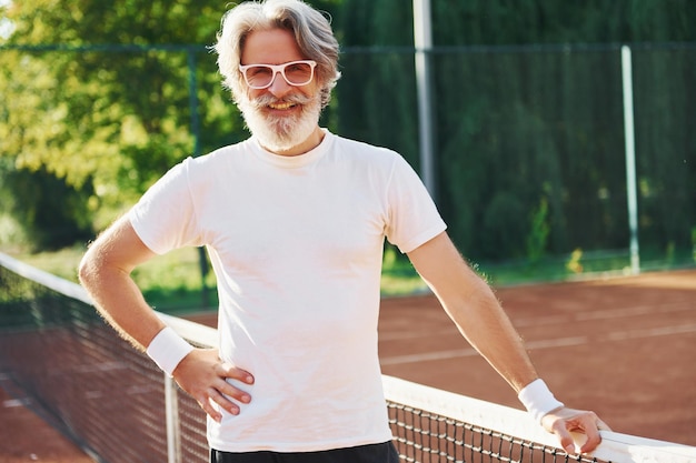 In posa per una macchina fotografica In occhiali da sole Senior uomo moderno ed elegante all'aperto sul campo sportivo durante il giorno