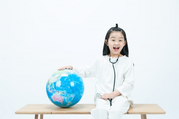 In un bianco, un bambino seduto su una sedia di legno sorride brillantemente con uno stetoscopio sul globo.