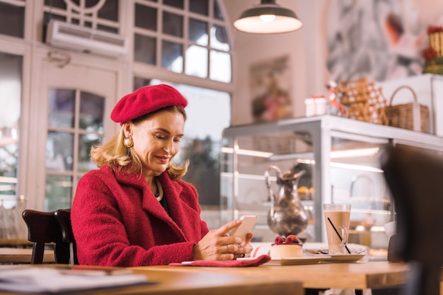 In una pasticceria accogliente. Bella donna matura che indossa accessori piacevoli e cappotto rosso in seduta accogliente panetteria