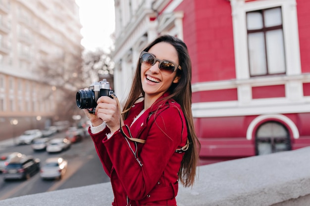 Incantevole ragazza con lunghi capelli castani che fotografa la città. Foto all'aperto di una bella fotografa in abiti rossi che si gode il fine settimana.