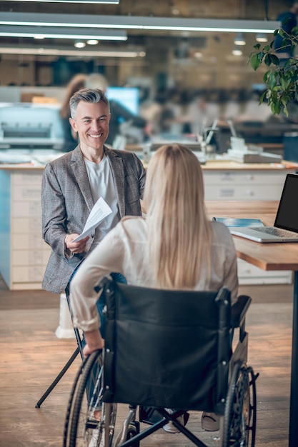 Inclusività. Uomo adulto sorridente in abbigliamento formale che porge documenti a una donna con lunghi capelli biondi su sedia a rotelle seduto con la schiena alla telecamera in ufficio