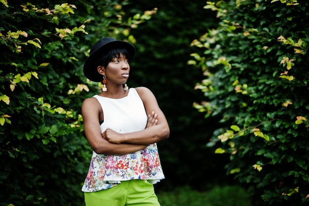 Incredibile donna modello afroamericano in pantaloni verdi e cappello nero in posa al parco