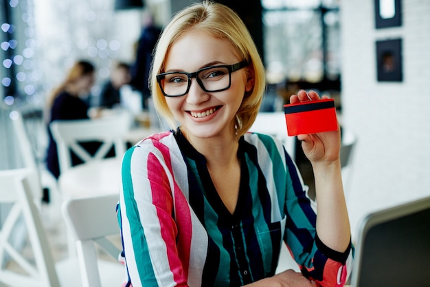 Incredibile ragazza con i capelli chiari che indossa camicia colorata e occhiali seduti in un caffè con laptop e carta di credito, concetto di freelance, shopping online, sorridente.