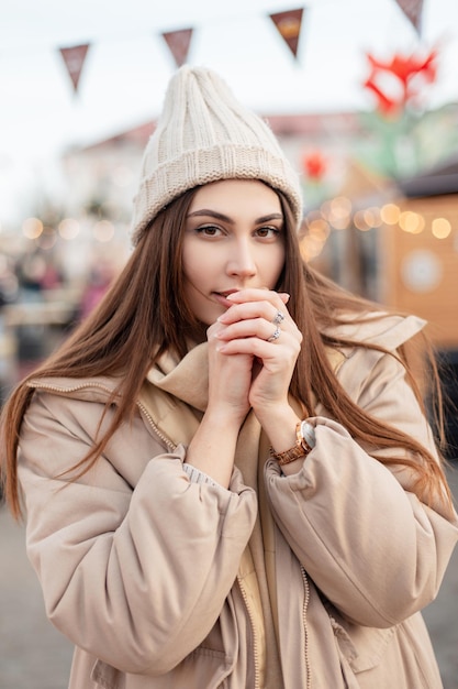 Incredibile ritratto di una bella ragazza alla moda in un capospalla beige con un cappello vintage lavorato a maglia, una giacca invernale e una sciarpa si trova in città e guarda la telecamera