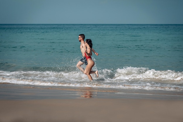 Incredibili sposini sulla spiaggia esotica che corrono gioiosamente nelle acque poco profonde, schizzi e risate intorno.