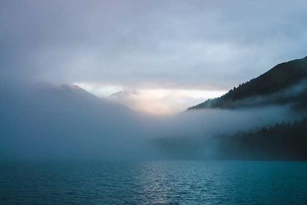 Increspature sulla superficie del lago di montagna nella nebbia nell'ora d'oro. Il sole splende attraverso dense nuvole basse nelle montagne. La traccia soleggiata brilla sull'acqua all'alba. Scenario alpino rilassante con nebbia dai toni pastello.