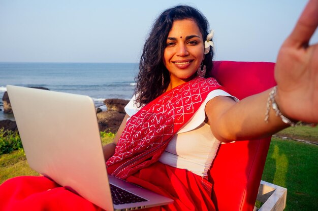 India donna freelancer asiatico fotografare selfie ritratto su smartphone donna in rosso indiano elegante saree sari lavorando su laptop lavoro remoto lavoro da sogno nella costa del paradiso