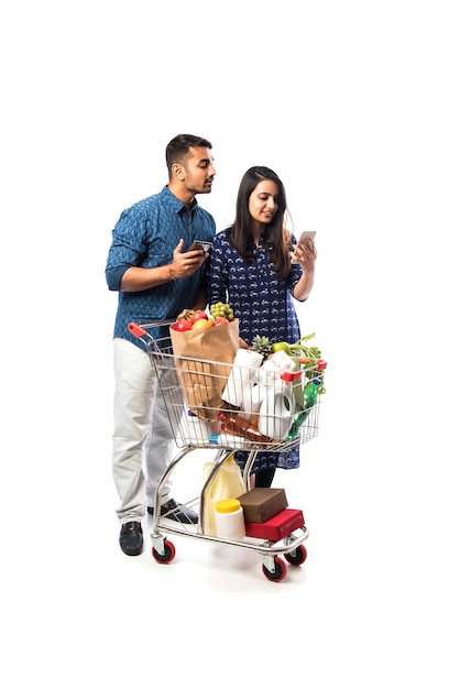 Indian giovane coppia con carrello o carrello pieno di generi alimentari, frutta e verdura. Isolato Foto a figura intera su muro bianco