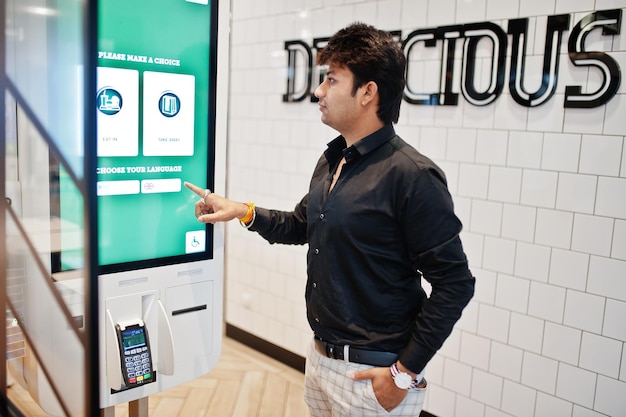 indian man customer at store place orders and pay through self pay floor kiosk for fast food payment terminal make a choise of language on screen