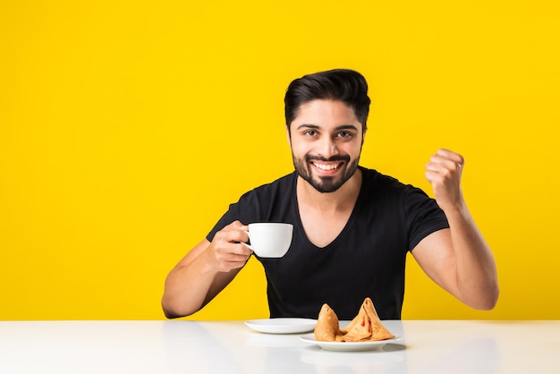 IndianÃ‚Â giovane in possesso di un Teacup, bere e godersi l'aroma del caffè. Ragazzo asiatico seduto al tavolo su sfondo giallo con tazza