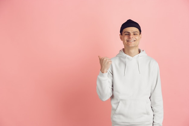 Indicando a lato. Il ritratto moderno del giovane caucasico isolato sul fondo rosa dello studio. Bellissimo modello maschile in bianco. Concetto di emozioni umane, espressione facciale, vendite, pubblicità. Copyspace.