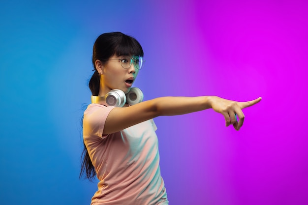 Indicare, mostrare. Ritratto di giovane donna asiatica su sfondo sfumato in neon. Bellissimo modello femminile in stile casual. Concetto di emozioni umane, espressione facciale, gioventù, vendite, pubblicità.