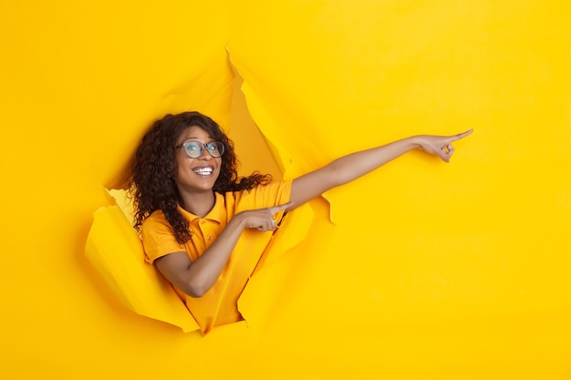 Indicazione felice pazza. Giovane donna afroamericana allegra in fondo di carta gialla strappata, emotiva, espressiva. Rompere, sfondare. Concetto di emozioni umane, espressione facciale, vendite, pubblicità.