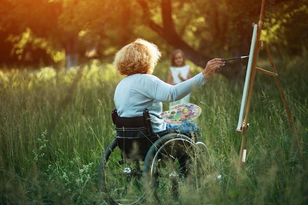 Indietro Colpo Di Donna Paraplegica Disegnare Sua Figlia Nel Parco. Donna Brillante Al Sole Dipinge Sul Cavalletto Mentre Era Seduto In Una Sedia A Rotelle.