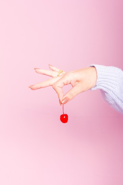 Indossa un maglione blu e tiene in mano una ciliegia. Sfondo rosa