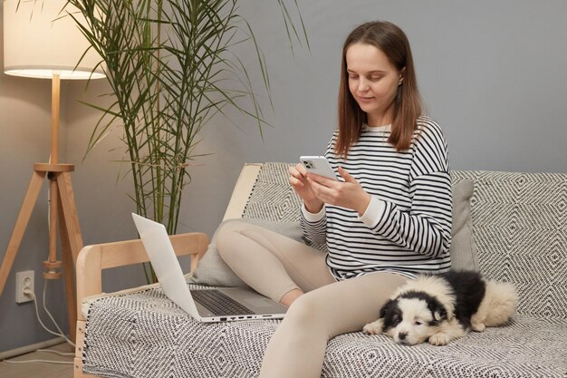 indossa una camicia a righe seduta sul divano con il suo cucciolo usando un computer portatile e uno smartphone