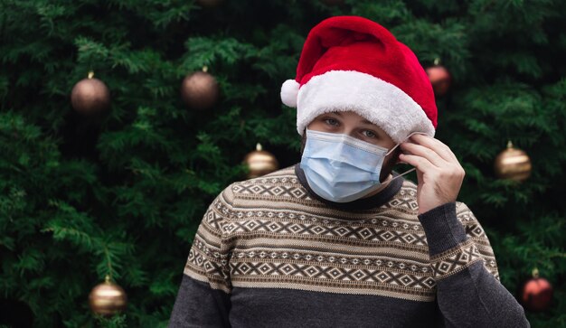 Indossare correttamente una maschera medica. Close up Ritratto di uomo che indossa un cappello di Babbo Natale, maglione di Natale e mascherina medica con emozione. Sullo sfondo di un albero di Natale. Pandemia di coronavirus