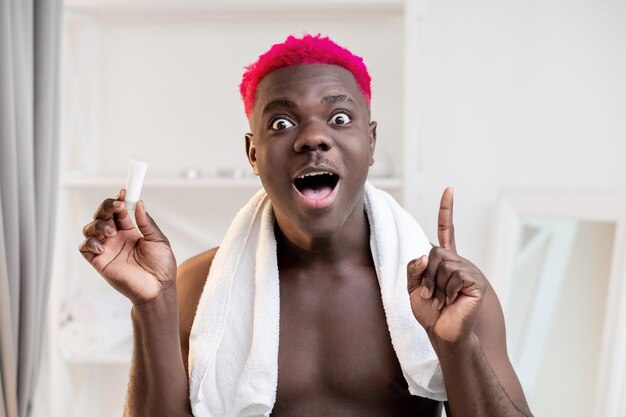 Indovina l'idea Uomo di colore sorpreso Cosmetici per la cura della pelle Trattamento perfetto Gesto di Eureka Ragazzo africano senza maglietta scioccato con capelli rosa brillante che tiene il dito in su tenendo l'interno del bagno in tubo di crema