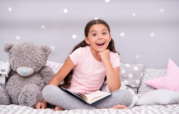 Indovina la sua fiaba preferita Bambina seduta a letto con orsacchiotto leggi il libro Bambino si prepara ad andare a letto Tempo piacevole nell'accogliente camera da letto Bambina bambino capelli lunghi carino pigiama rilassarsi e leggere il libro per sopportare il giocattolo